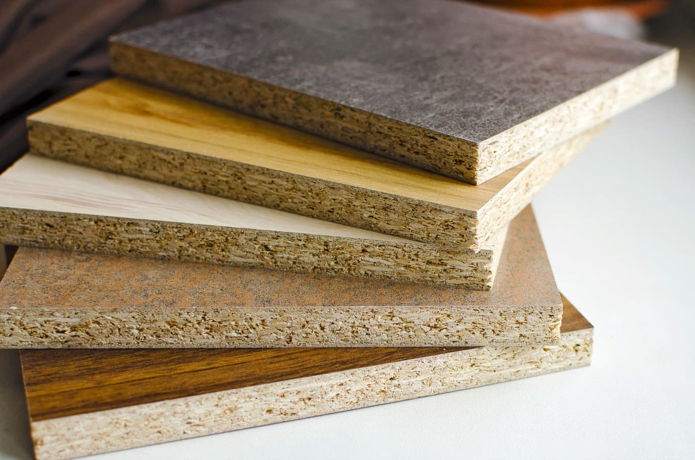 Une pile de panneaux mélaminés vus de trois quarts : les faces portent des décors bois et pierre, tandis que les tranches laissent voir l’aggloméré brut, fait de copeaux de bois collés.